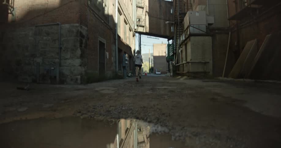 A woman is running down a narrow dimly lit alleyway with a large puddle in the foreground reflecting the surrounding buildings and the runner