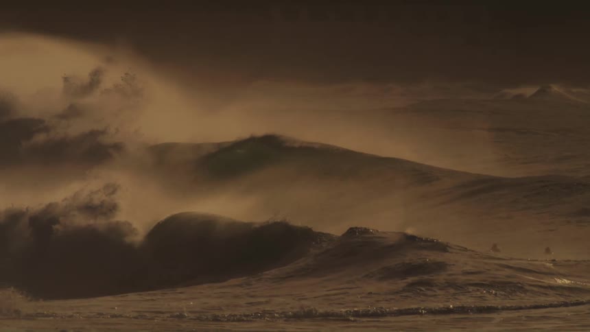 Waves crash into the sea at sunset with foam being blown by the wind as surfers wait in the water
