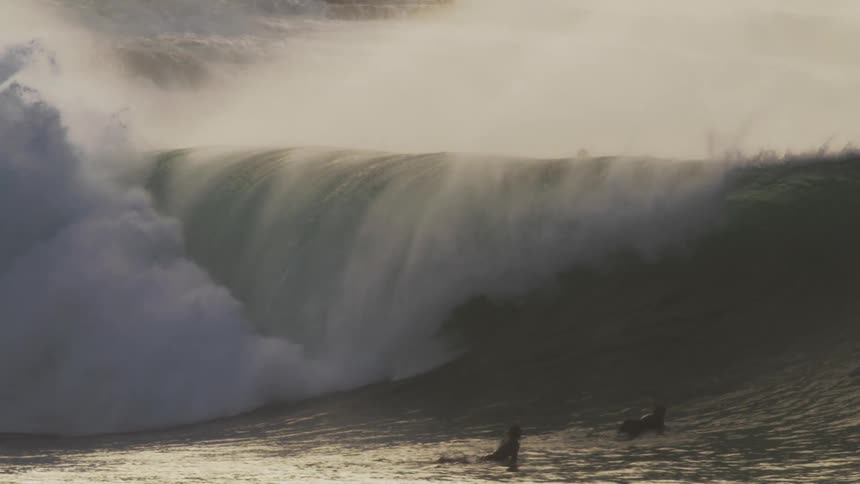 A massive wave crashes in a swirl of foam and spray with surfers dwarfed by the size of the wave