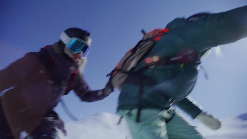 Two individuals in winter gear are preparing to board a helicopter with a logo on its side