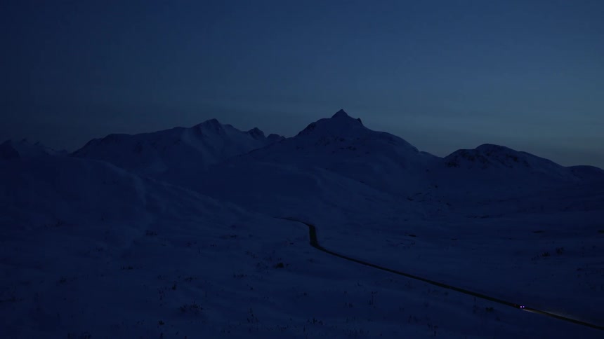 A dark twilight scene of a snowcovered mountainous landscape with a winding road leading into the distance and a faint light from a vehicle visible on the road