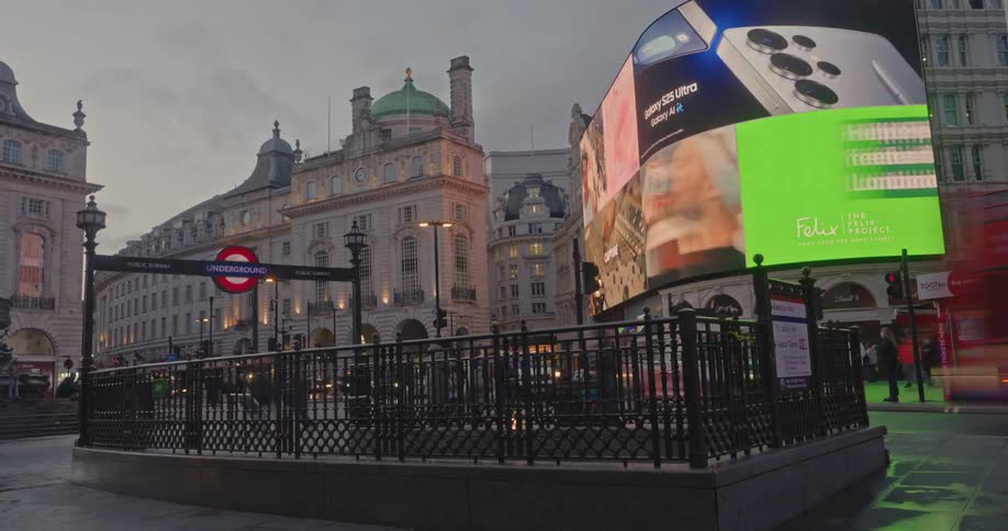 Red doubledecker buses and pedestrians pass by the iconic curved advertising screens and historic buildings of piccadilly circus in london under an overcast sky
