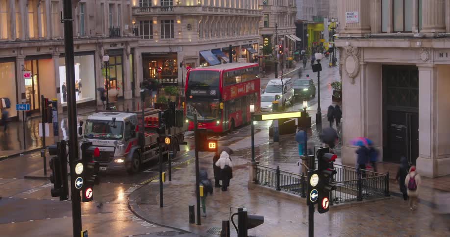 Pedestrians walk along a sidewalk downtown and cross a busy intersection on a rainy day