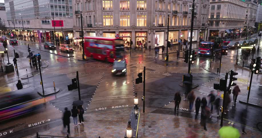 Pedestrians and vehicular traffic cross a busy intersection near brightly lit stores while it rains