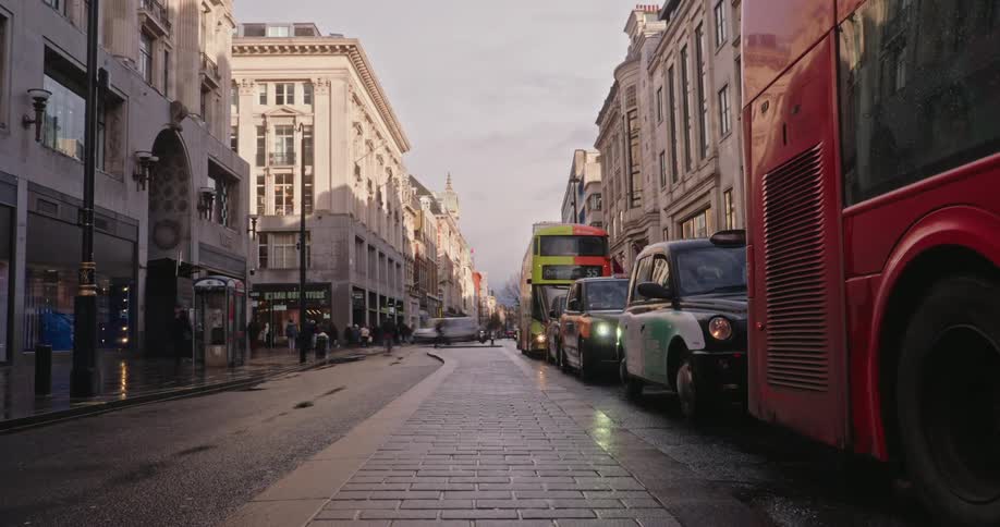 Busy london street with red buses and black taxis lined up alongside the wet pavement reflecting the soft evening light between historic buildings