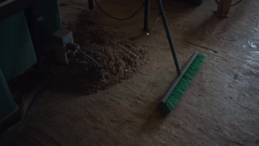 A green broom leans against a table saw in a woodworking shop on a wooden floor with sawdust