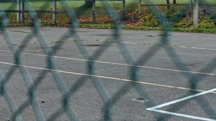A cracked asphalt playing surface with worn lines is seen through a chain link fence