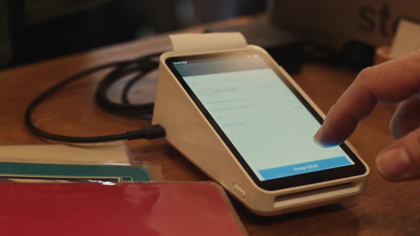 Person types on a touchscreen payment terminal placed on a wooden counter and another makes a contactless payment with a card