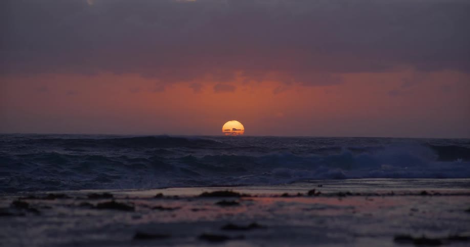 A bright orange sun sets over an ocean beach against a pink sky