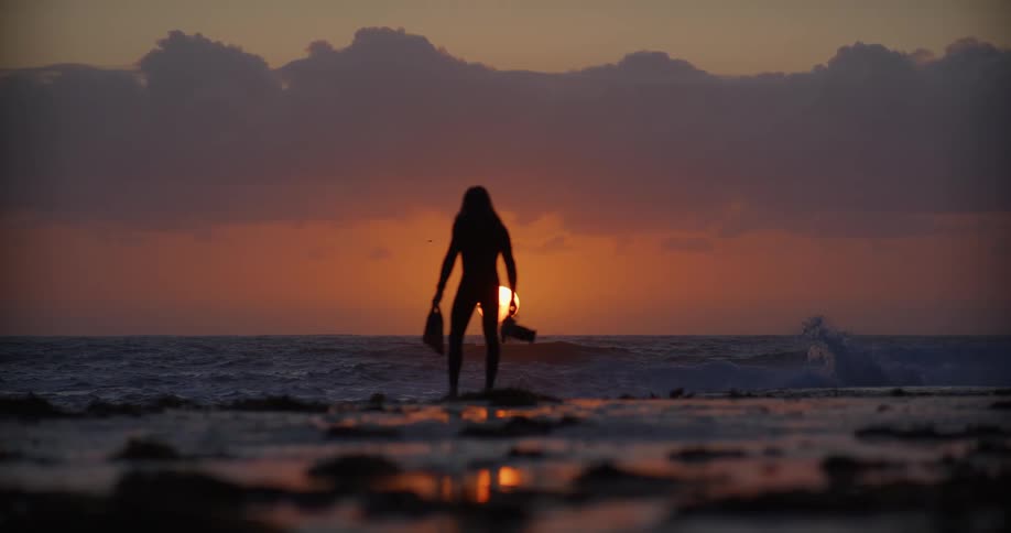 A silhouetted man stands on the beach at dusk holding foot fins in one hand and an underwater camera in the other as turbulent waves crash ashore