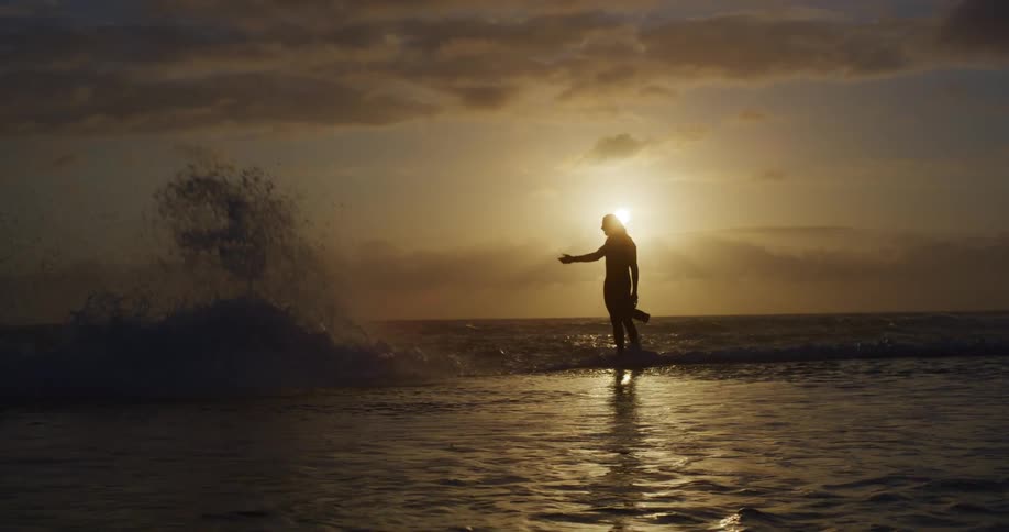 A man holding a camera stands in the ocean and puts his arm out as if he is creating a huge wave