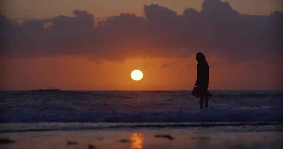 Male photographer standing in the surf silhouetted against the sun sitting near the horizon holds a camera and flippers