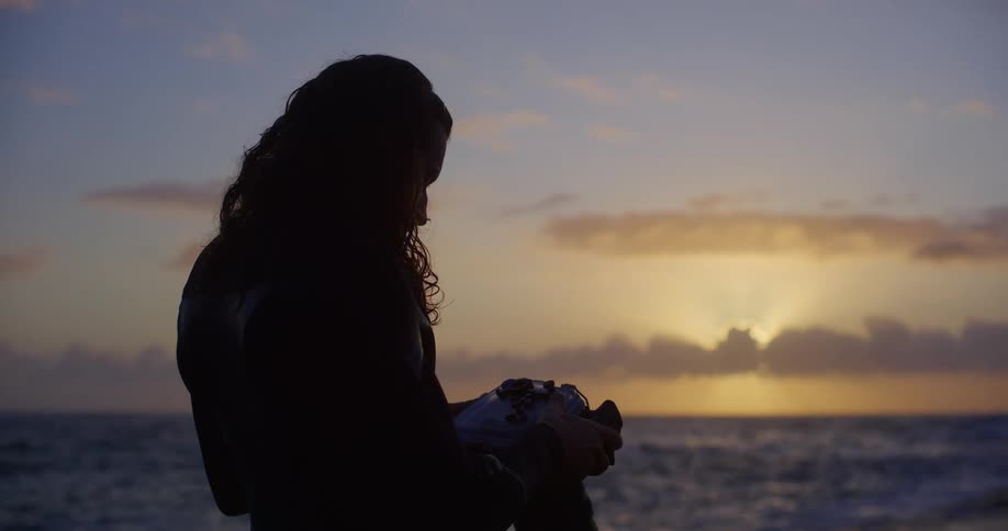 Male photographer wearing a wet suit examines his camera while standing on the shore at sunset