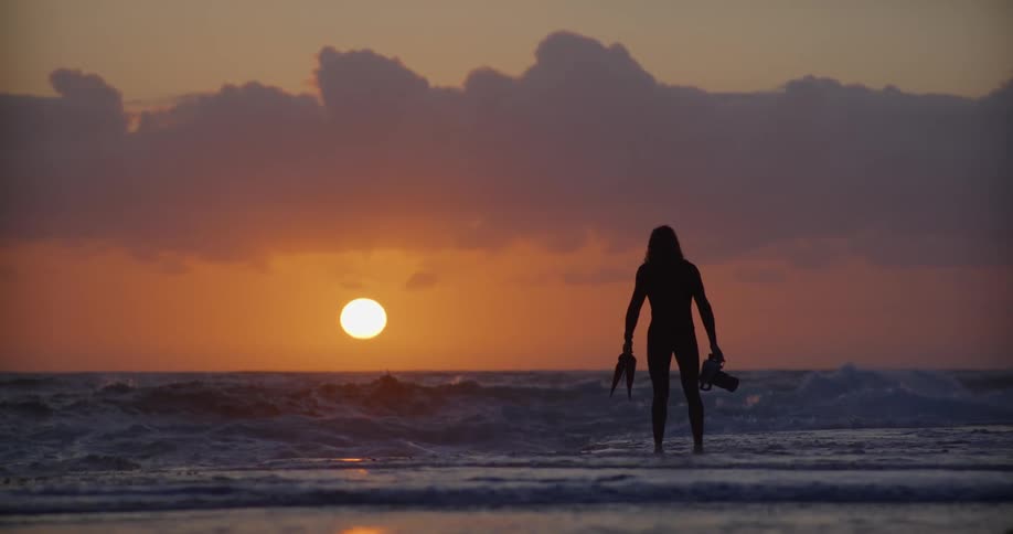 A diver in silhouette form is holding a camera and walking into the ocean