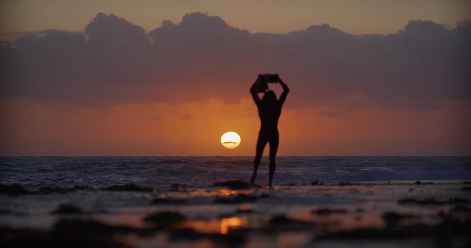 A silhouetted figure stands on a beach at sunset holding what appears to be fins and a camera with a sun sinking behind the ocean horizon and clouds in the background