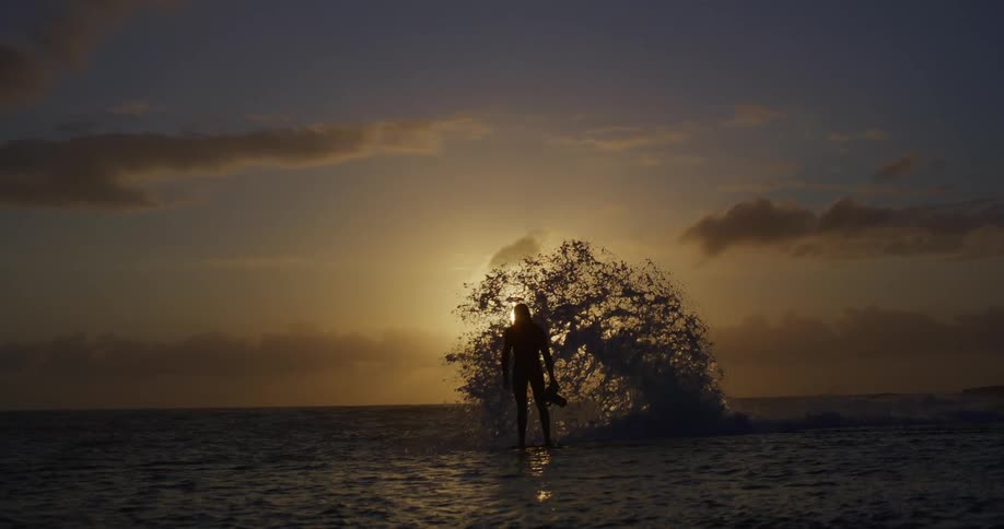The silhouette of a person standing in shallow water and then a wave splashing in front of them