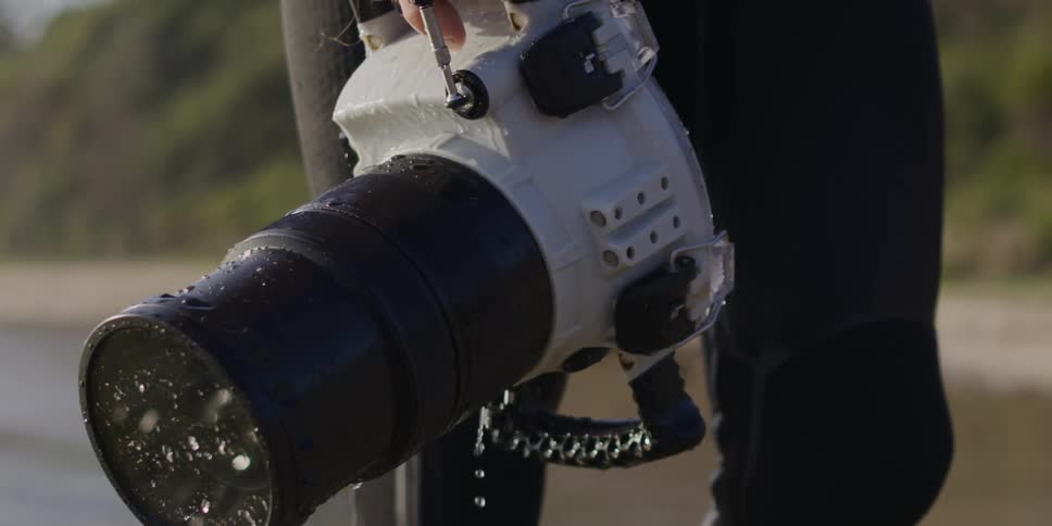 A person in a wetsuit holds a dripping underwater camera while walking along the coast with a hill in the background