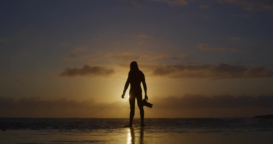 A silhouetted figure stands on the beach raising a camera from one hand to both hands as the sun sets behind them