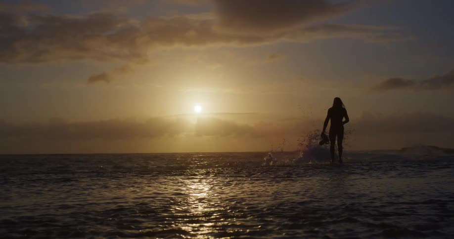 A silhouetted figure wades through shallow water at sunset holding an underwater camera in one hand as the sun dips below the horizon