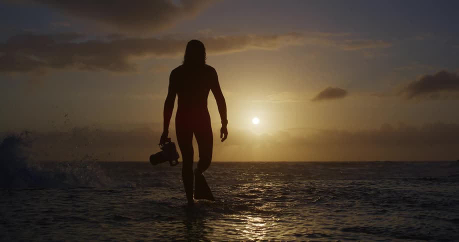 A mans silhouette is shown walking forward in the ocean holding a camera