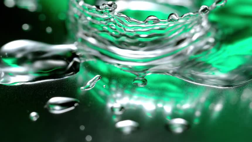 A water droplet disturbs a calm water surface falling onto a puddle on a reflective green surface creating ripples