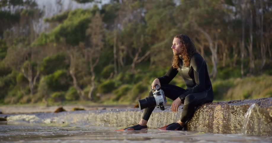 Male photographer wearing a wet suit sits on a ramp near shallow water on an autumn day
