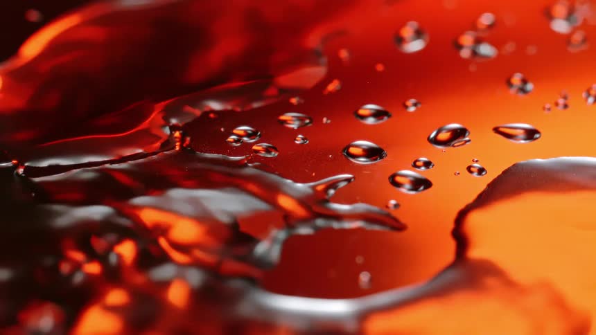 A water drop falls into a puddle on a reflective orange surface creating ripples and reflecting the drop