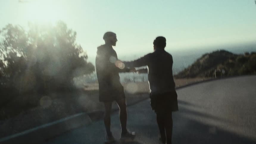 Two friends walk along a coastal road wearing shorts and sweatshirts on a sunny day