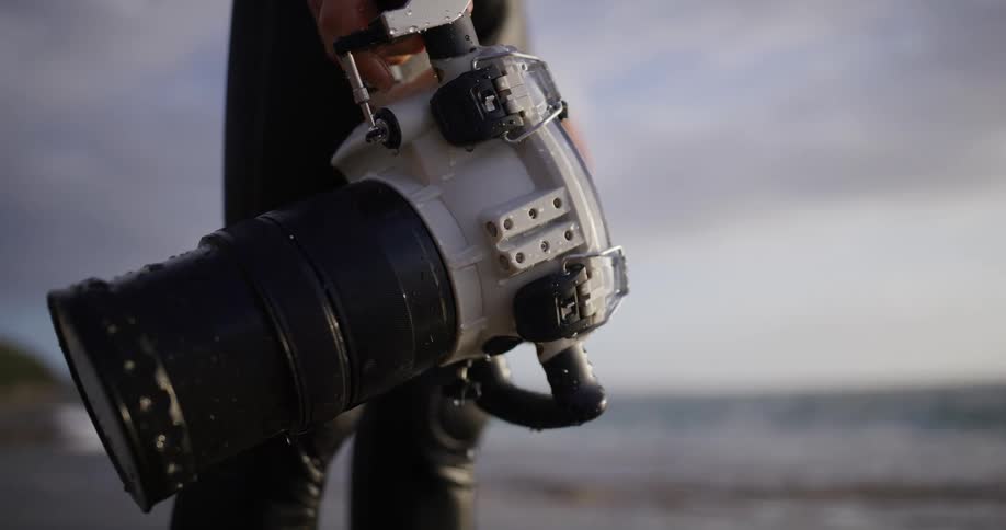 A closeup captures an underwater camera dripping with water as a person holds it on the beach with waves softly out of focus in the background