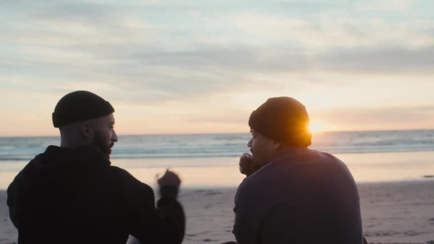 Two male friends sitting enjoying the sunrise on the horizon and clapping their hands