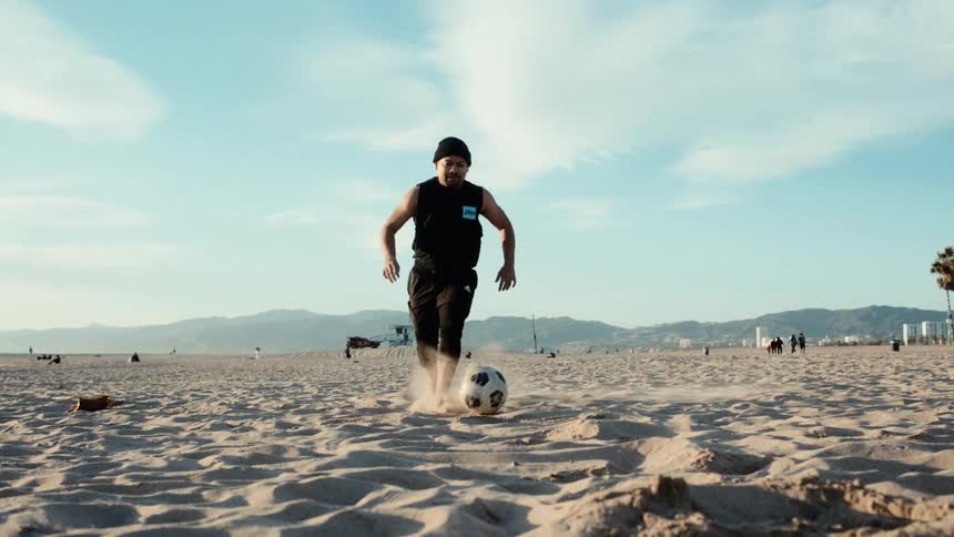 A hispanic man plays soccer on the beach on a partly cloudy day
