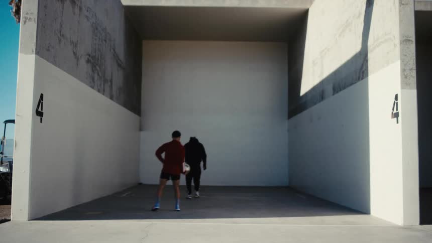 Two men in athletic attire energetically throw and kick a soccer ball inside a racquetball court labeled 4 practicing ballhandling skills and footwork in the confined space