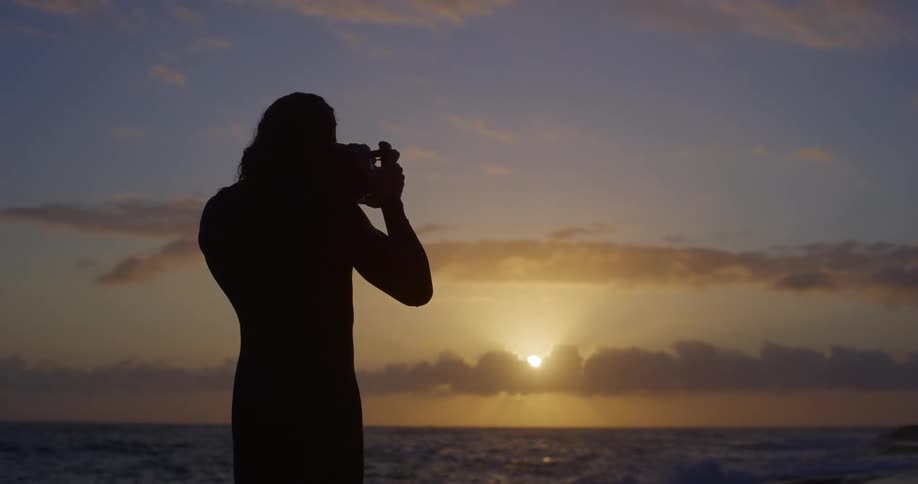 A silhouetted figure with long hair stands at the oceans edge holding a camera and looking at it against a vibrant sunset sky