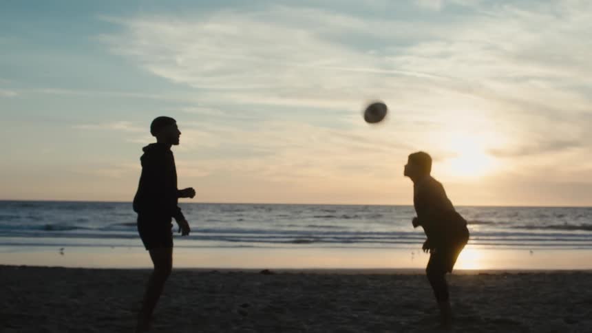 Silhouetted figures play with a ball on a beach at sunset