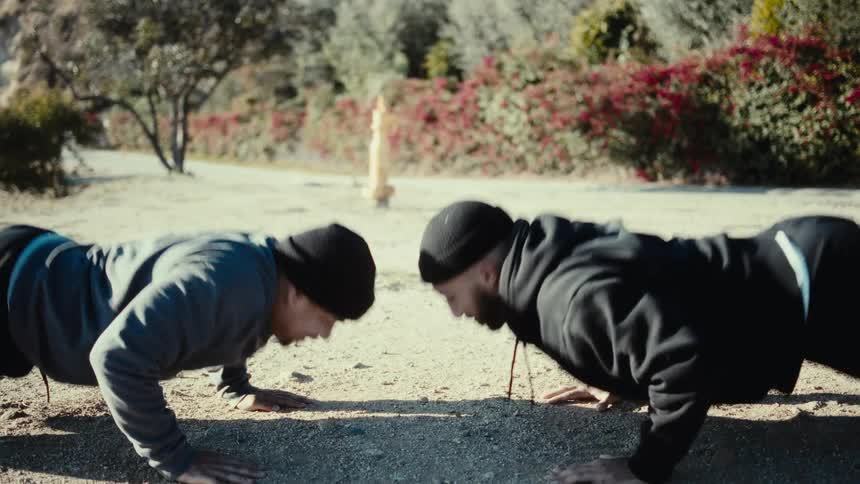 Two men in beanies one in a black hoodie and the other in grey do pushups facetoface and touch hands midexercise