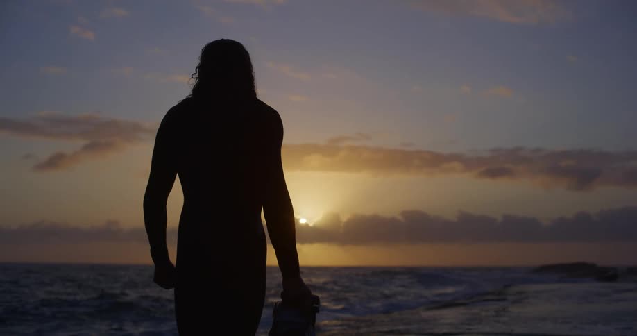 The silhouette of a man stands at the shoreline holding a camera and looking into the ocean