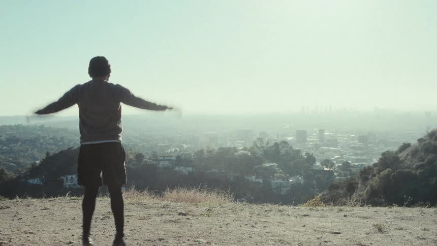 A person does jumping jacks on a hillside overlook with the city spread out below on the horizon