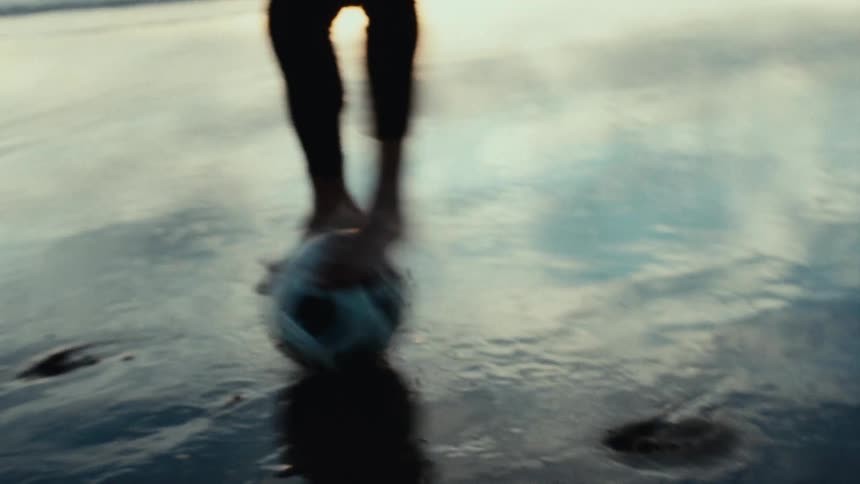 Two people play soccer on a wet sandy beach at sunrise dribbling and leaving footprints as they go
