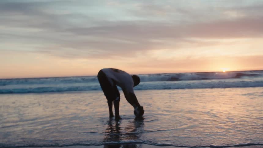 A man plays with a soccer ball on the beach after washing it in shallow ocean water at sunset