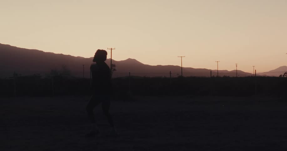 A silhouetted figure juggles a soccer ball on a dusty field at dusk with mountains and power lines in the background