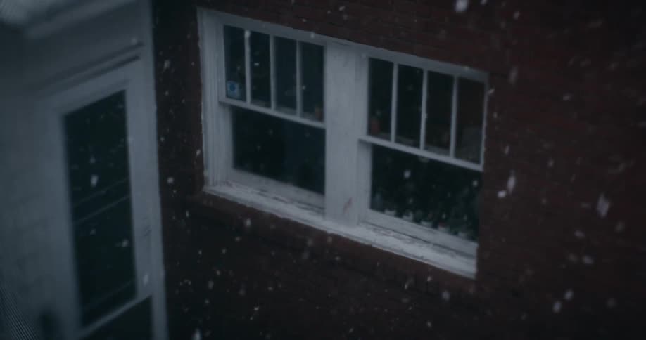 Snow falls softly outside a window viewed from a higher angle with a brick building visible