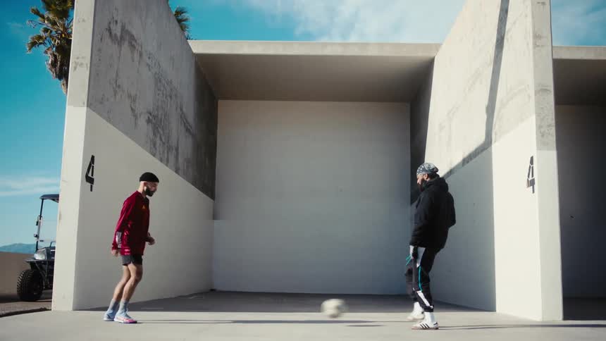 Two men one in black and one in red kick a soccer ball back and forth in front of a handballracquetball court with a partial golf cart on the left