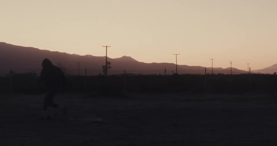 A person is silhouetted against a sunset sky jumping and stretching in preparation for exercise on a dirt field with mountains and power lines in the background