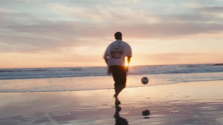 A person kicks a soccer ball on a beach at sunset with waves coming ashore in the background