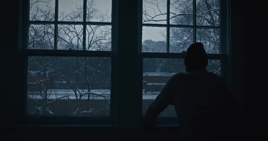 Silhouette person looking out window in a dark room watching it snow