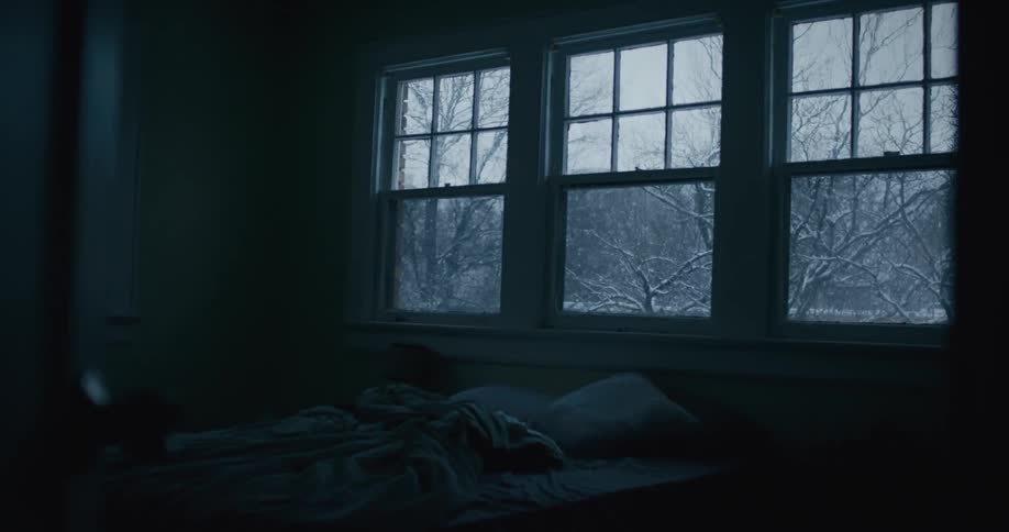 A view of a bedroom with a messy bed and three windows on the wall that look out onto a snowy outside area