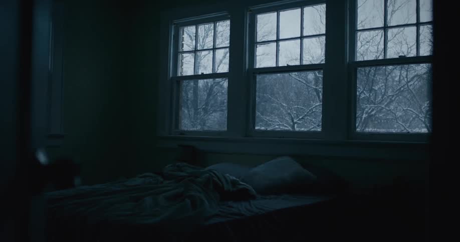 A view of a bedroom with a messy bed and three windows on the wall with a view of a blizzard outside