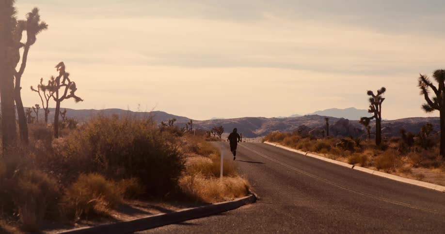 A person runs on a desert road surrounded by joshua trees under a pale sky
