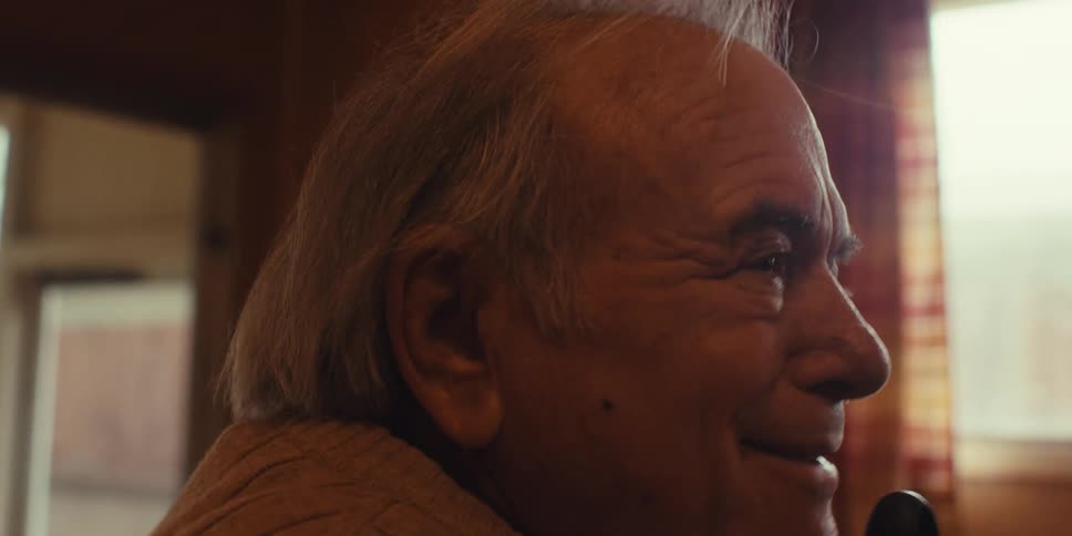 An older man in a sweater sings into the end of a broom handle smiling and moving his hand in time with the music