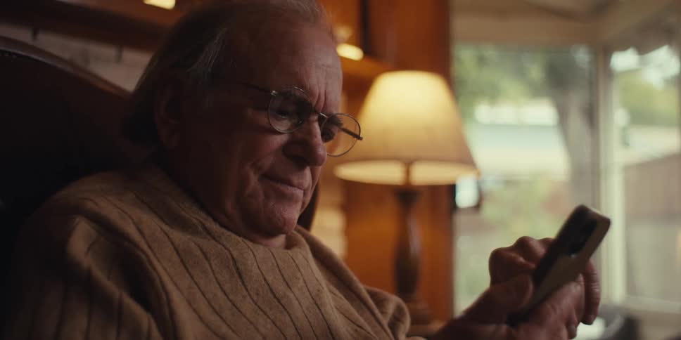 An elderly man is sitting in a cozy room at home browsing a smartphone